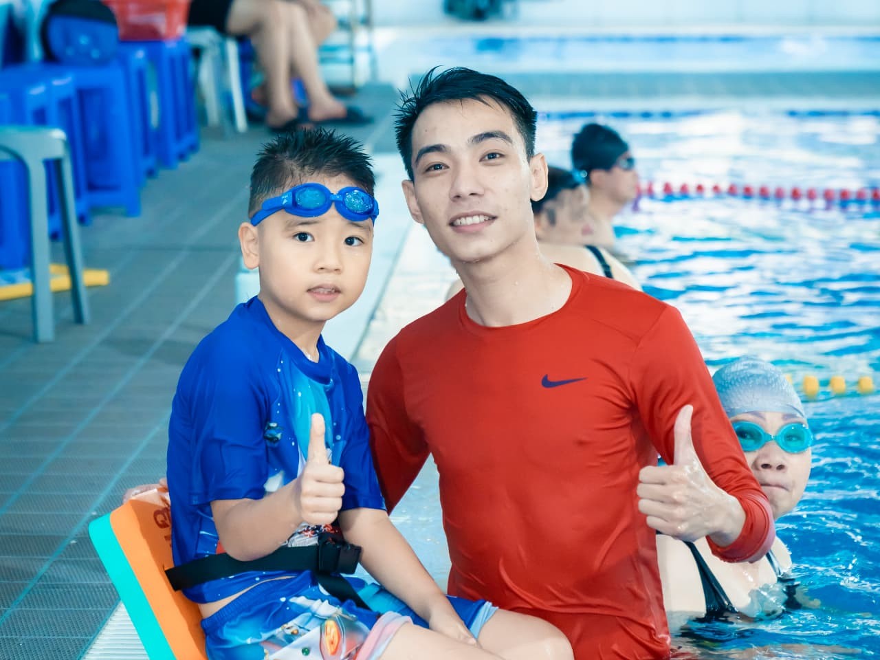 huấn luyện viên và một bé trai giơ tay khen tại mép bể bơi trong nhà